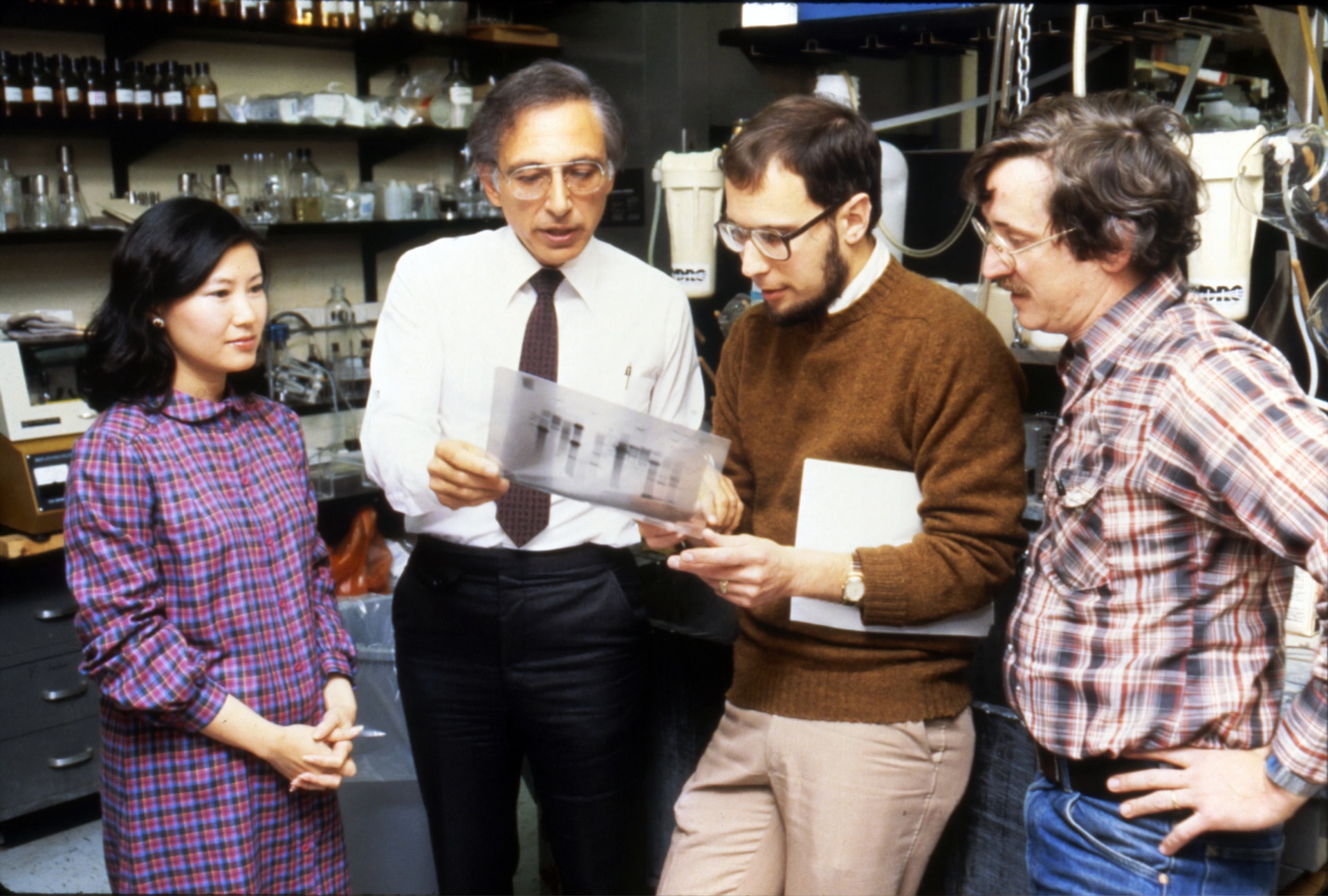 Four people dressed in late 1970s / early 1980s business casual clothes look at a transparent sheet with markings on it