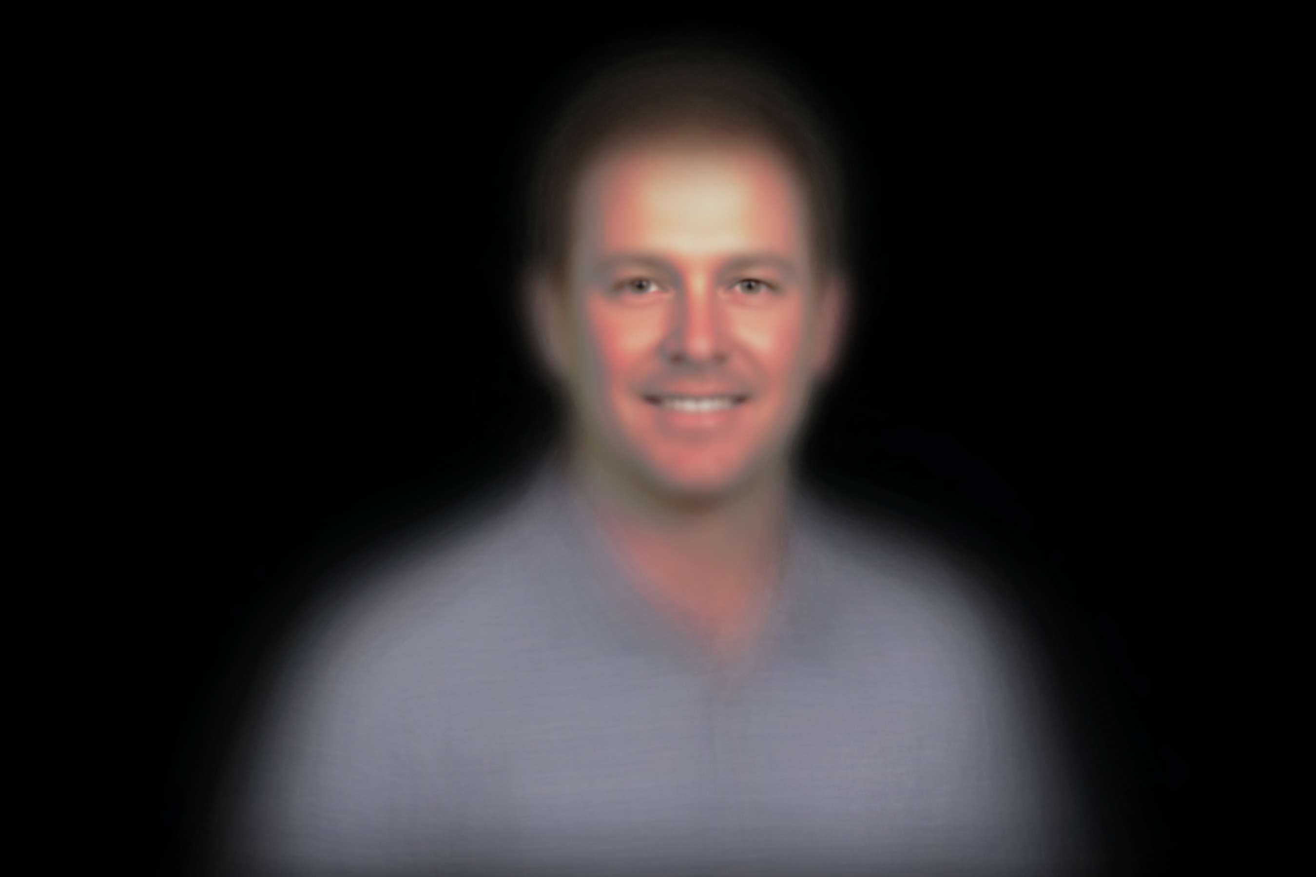 Generated image of a generic middle-aged white man in front of a balck background looking at the camera. His eyes are in sharp focus, other parts of his face are blurry, and everything besides his face is very blurry.