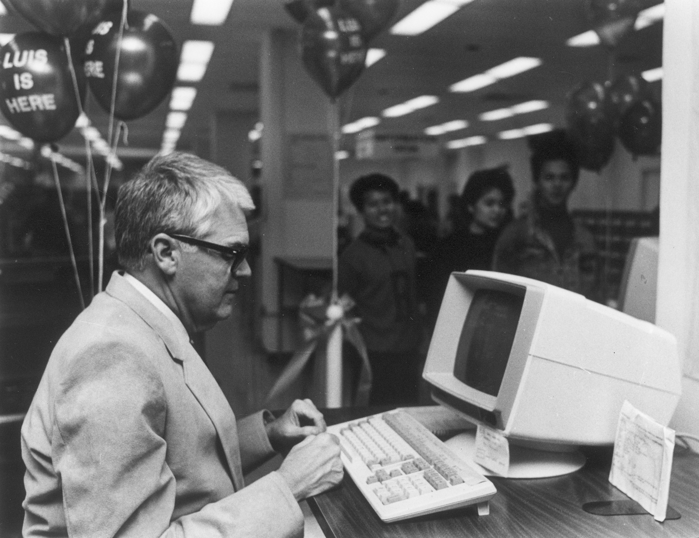 A black and white photo of a grey-haired person sitting in front of a 1970s-style computer terminal. In the background, other people look on.