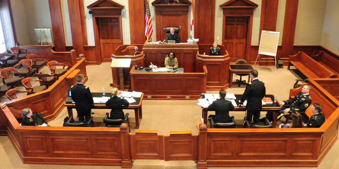 A wide, high-angle shot of an ornate courtroom. A judge and stenographer sit in the middle, with a witness in military garb in the witness box. In front of them there are two tables with prosecutors, defence attourneys, and others in military garb.