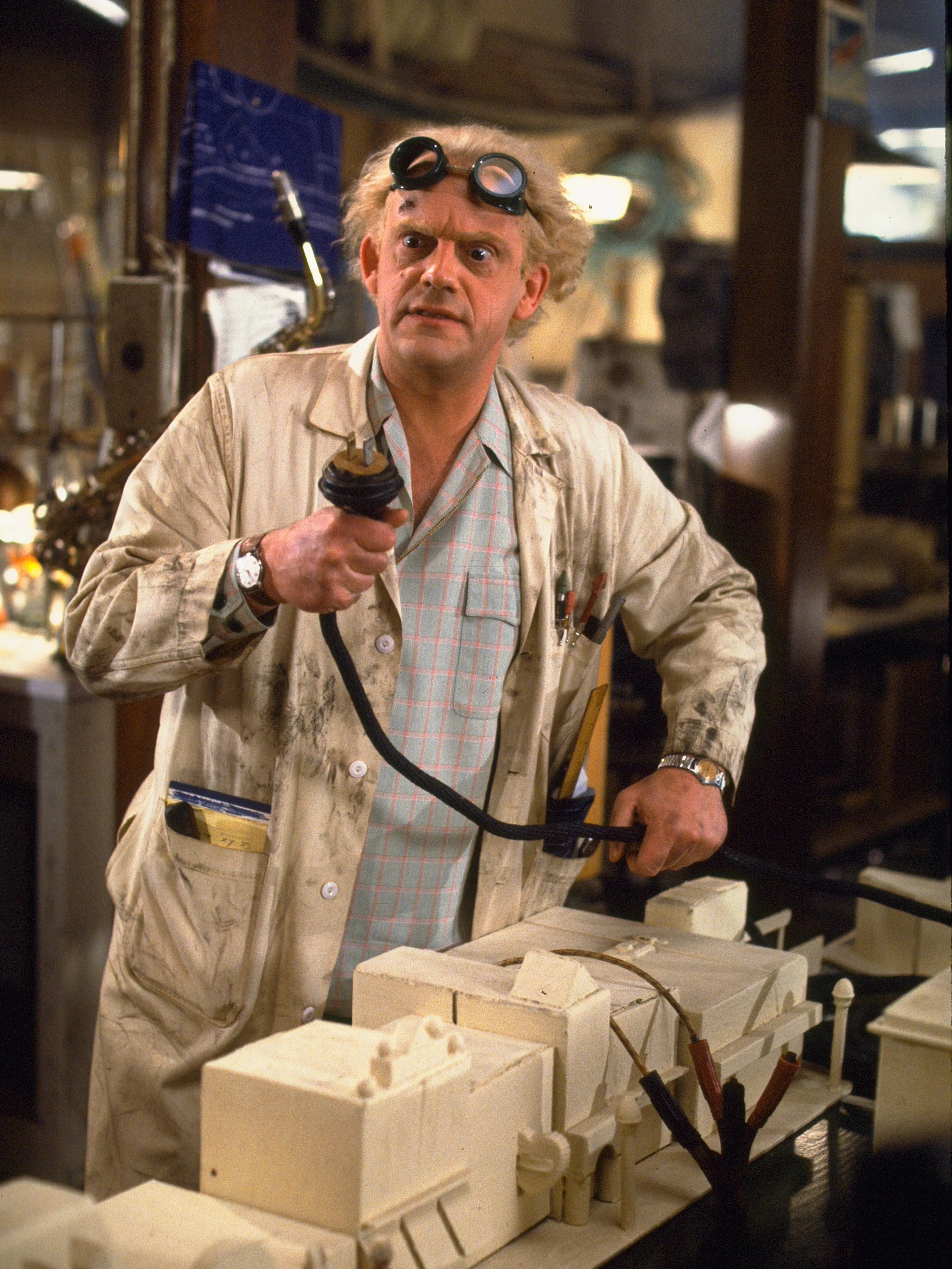 A man with wild white hair wears a filthy lab coat and goggles on his forehead and hold a large cable in his hand. He is surrounded by inscrutible scientific tools. (Christopher Lloyd playing Doc Brown in Back to the Future (1985))