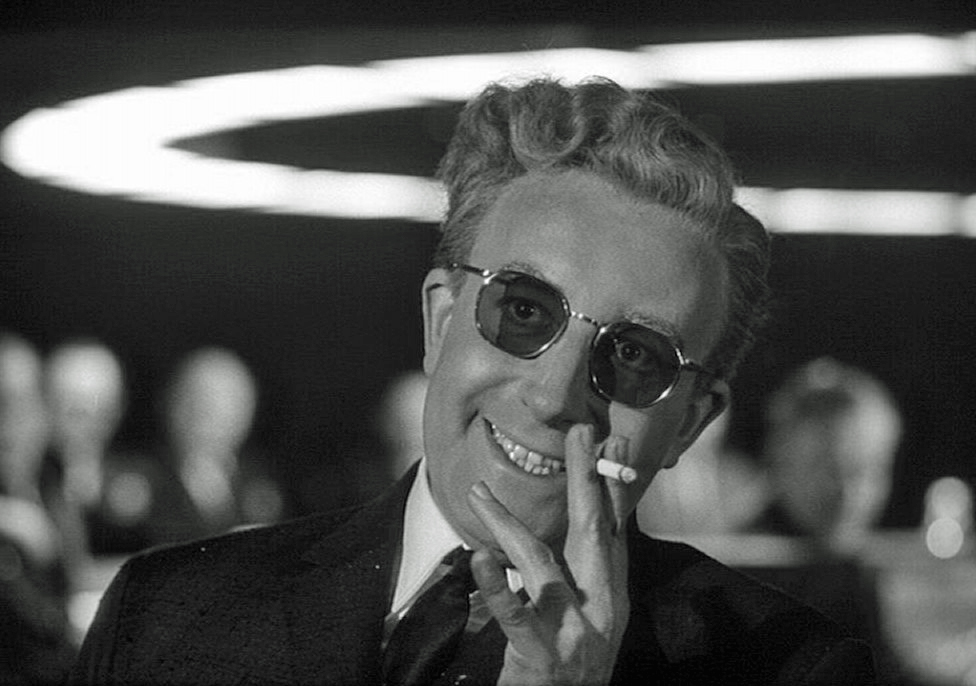 Still from Dr Strangelove (1964). Black and white image of a man wearing dark glasses with an unhinged grin on his face. He holds a cigarette near his face with one hand, with fingers held stiffly in odd directions.