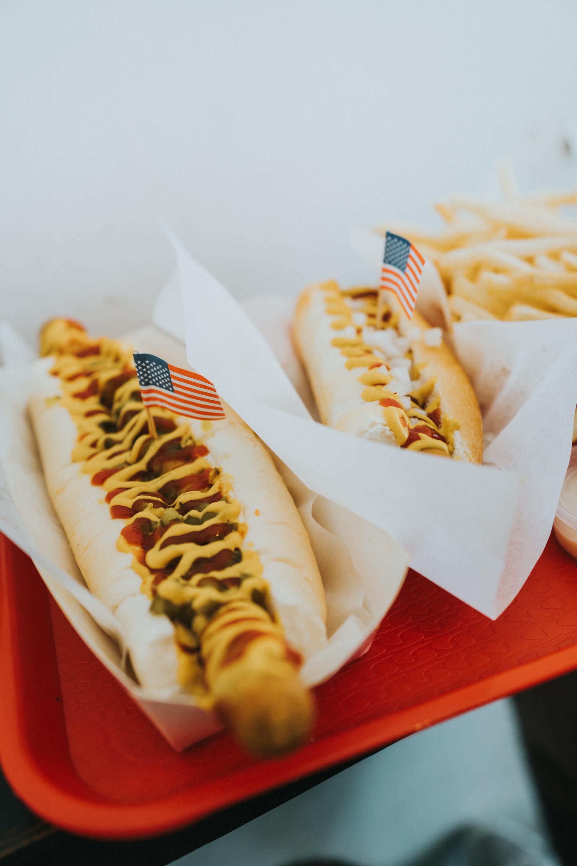 A photograph of a red tray with two long hotdogs, covered in ketchup and yellow mustard, with tiny American Flags stuck into them.