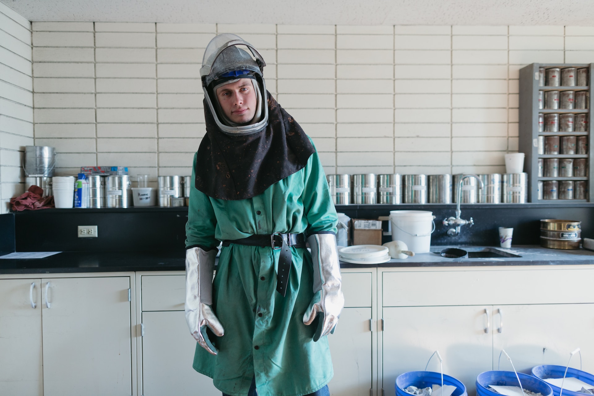 A man in full protective gear, including big silver glvoes and a helmet and a greeen  plastic coat, standing in front of some home-made lab equipment