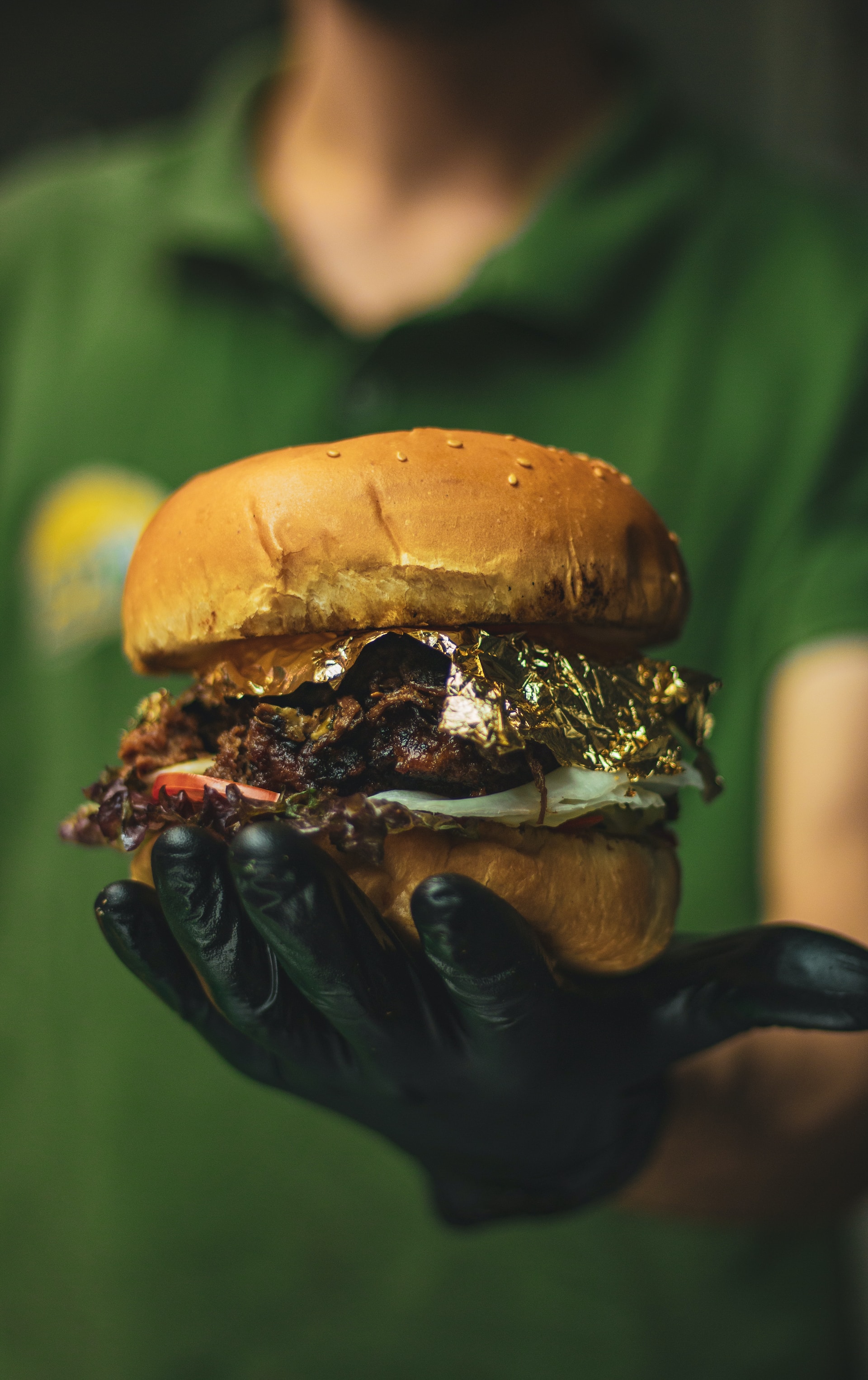 a person wearing a black disposable nitrile glove holding a hamburger with consipcuous gold leaf on top of the patty
