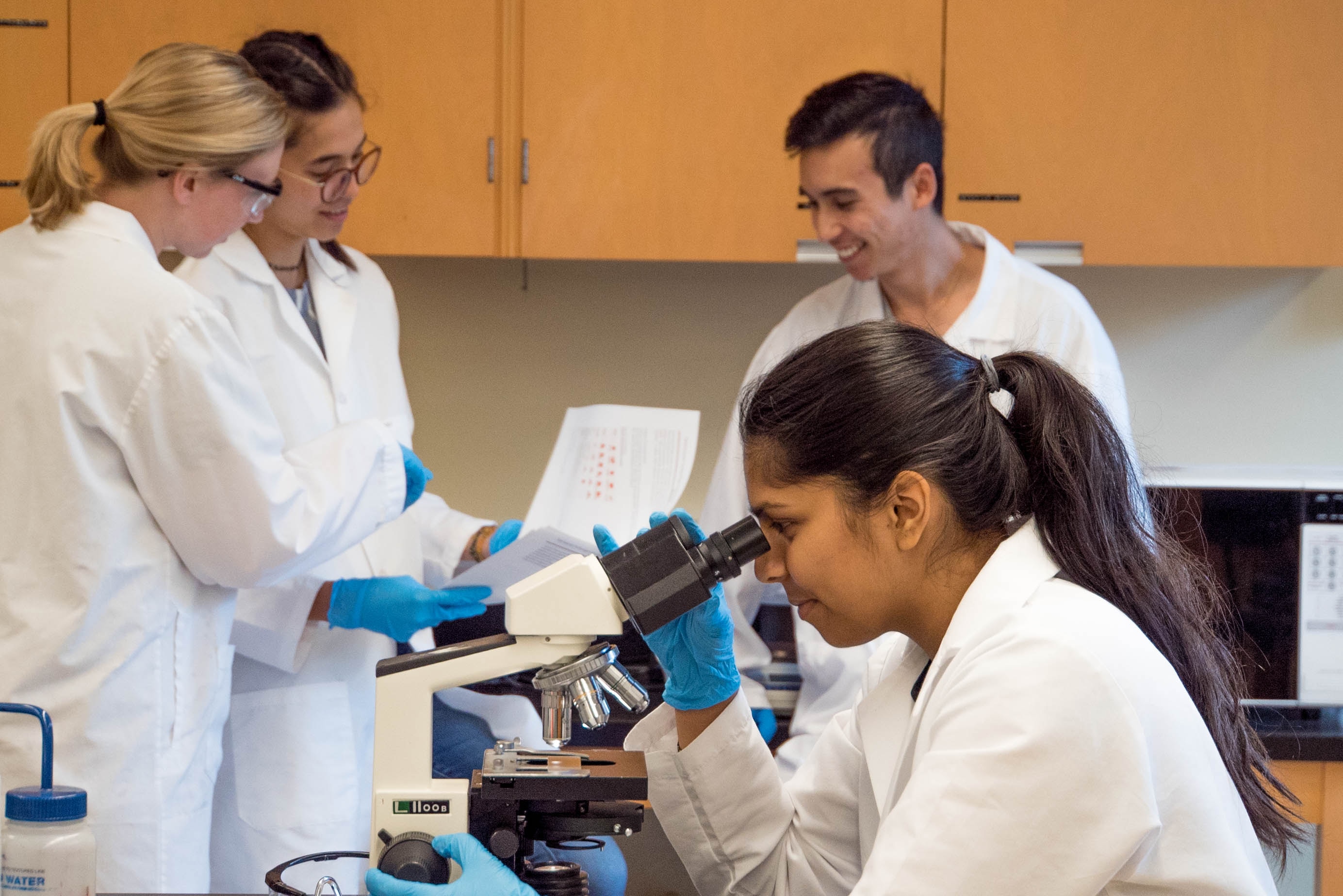 Four people in a scientific lab. One is looking through a microscope. The other three are smiling while looking at a paper document together.