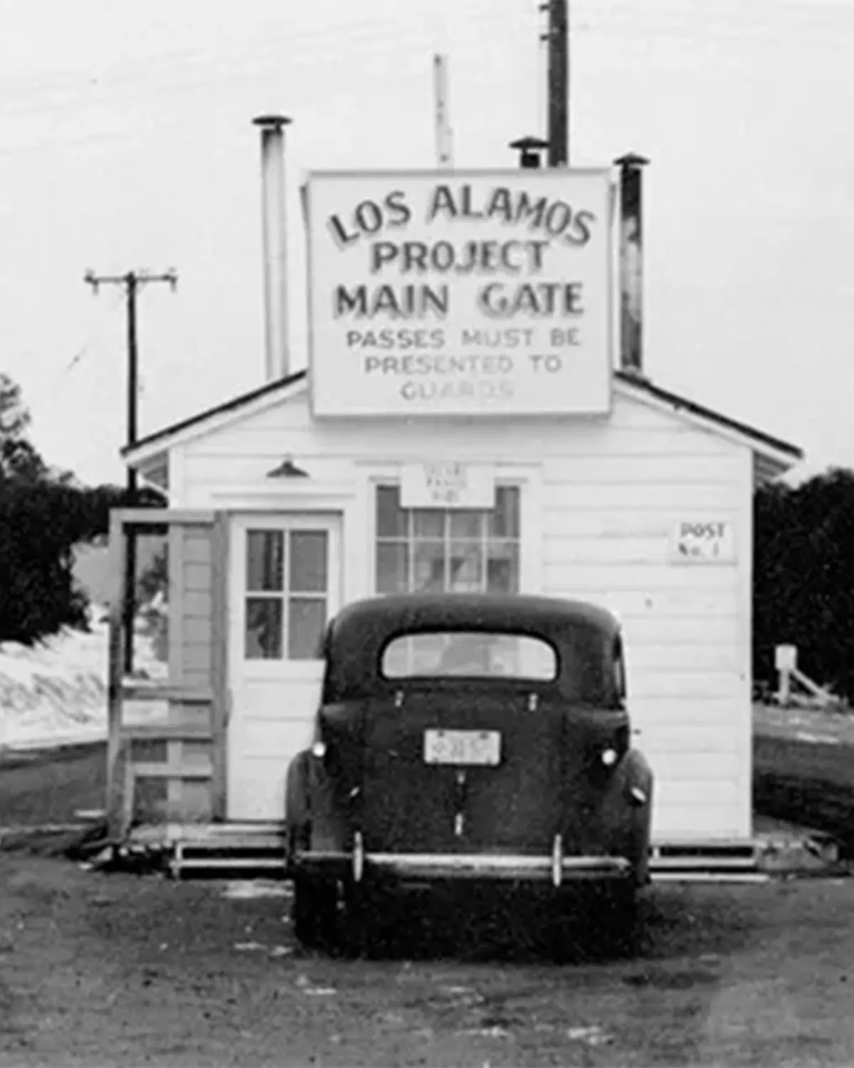 black and white photo of a small, white, wooden building with a 1940s-style car parked in front. A large sign on the front of the building reads 'LOS ALAMOS PROJECT MAIN GATE / PASSES MUST BE PRESENTED TO GUARDS'