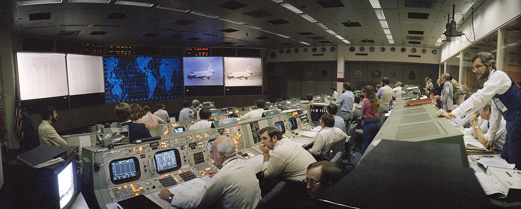 Photograph of NASA control room in the 1980s. A large room with big screens and rows of engineers and scientists sitting in front of computers