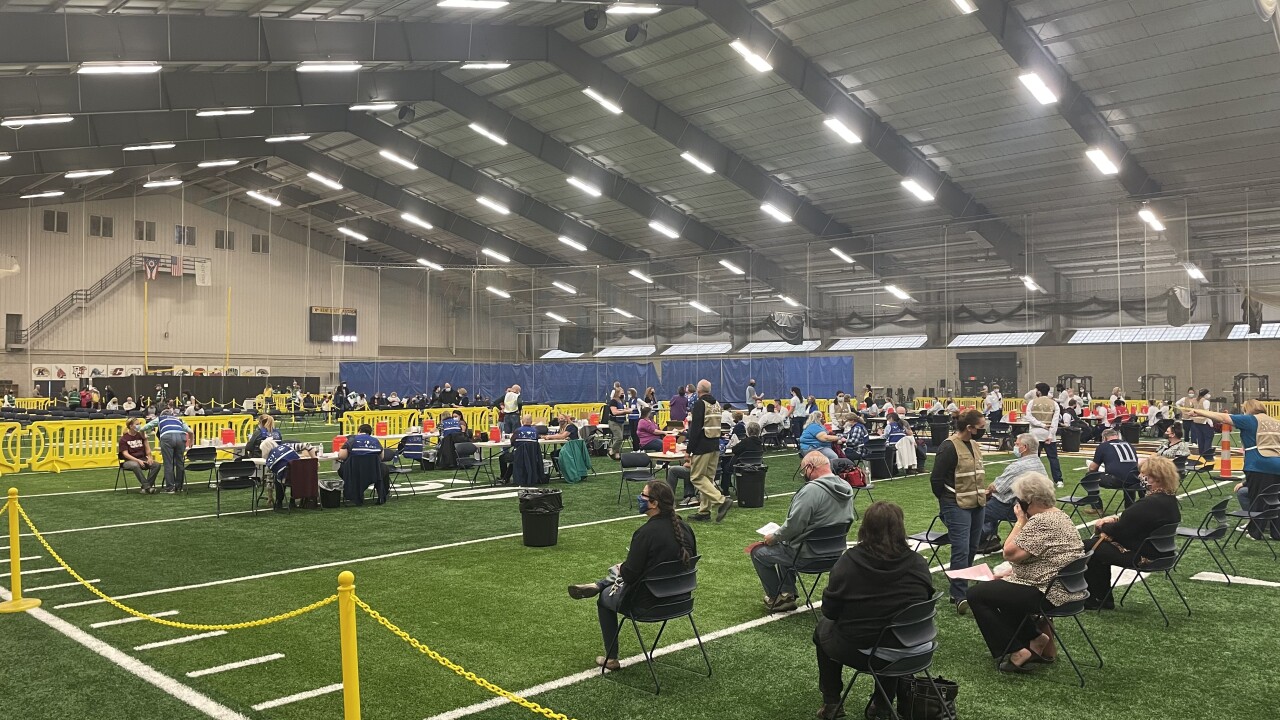 A crowd of people sitting in spaced chairs on the astroturf of an indoor stadium, waiting after receiving a vaccine.