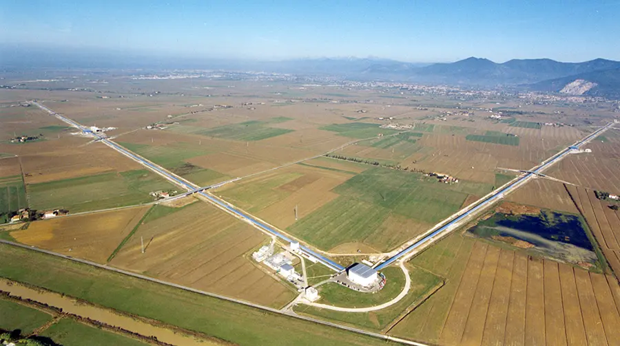 aerial photo of one site of a gravitational wave detector. There is a central hub building, from which extend two very long (3 km) arms at near-right angle from each other. The building is surrouned by what looks like farm land.