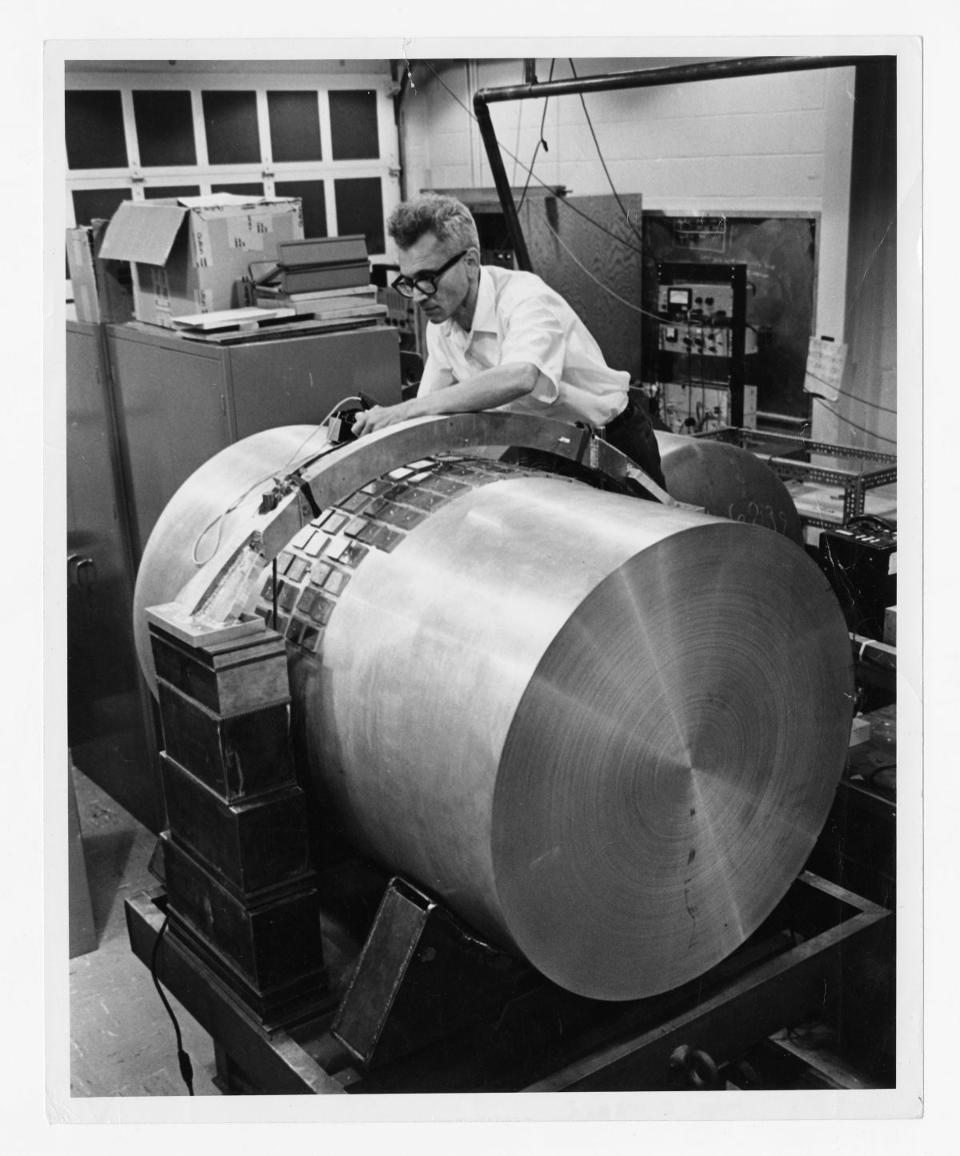 A black and white photo of a grey-haired scientist with sleeves rolled up leaning over a large metalic cylendar with complicating-looking electrical elements attached to it.