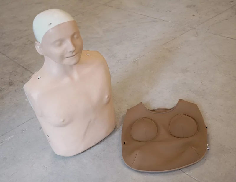 A photograph of a standard CPR dummy torso, which has a male physiology. Next to it is a neoprene vest featuring two 'breasts'.
